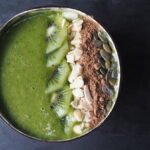 Close up picture of a green smoothie in a coconut bowl topped with slices of kiwi, flaked almonds, cacoa powder and pumpkin seeds