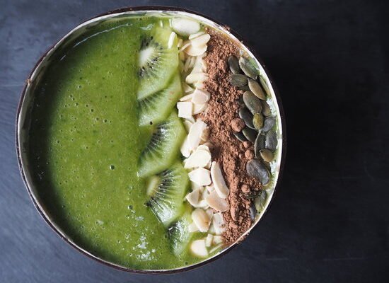 Close up picture of a green smoothie in a coconut bowl topped with slices of kiwi, flaked almonds, cacoa powder and pumpkin seeds